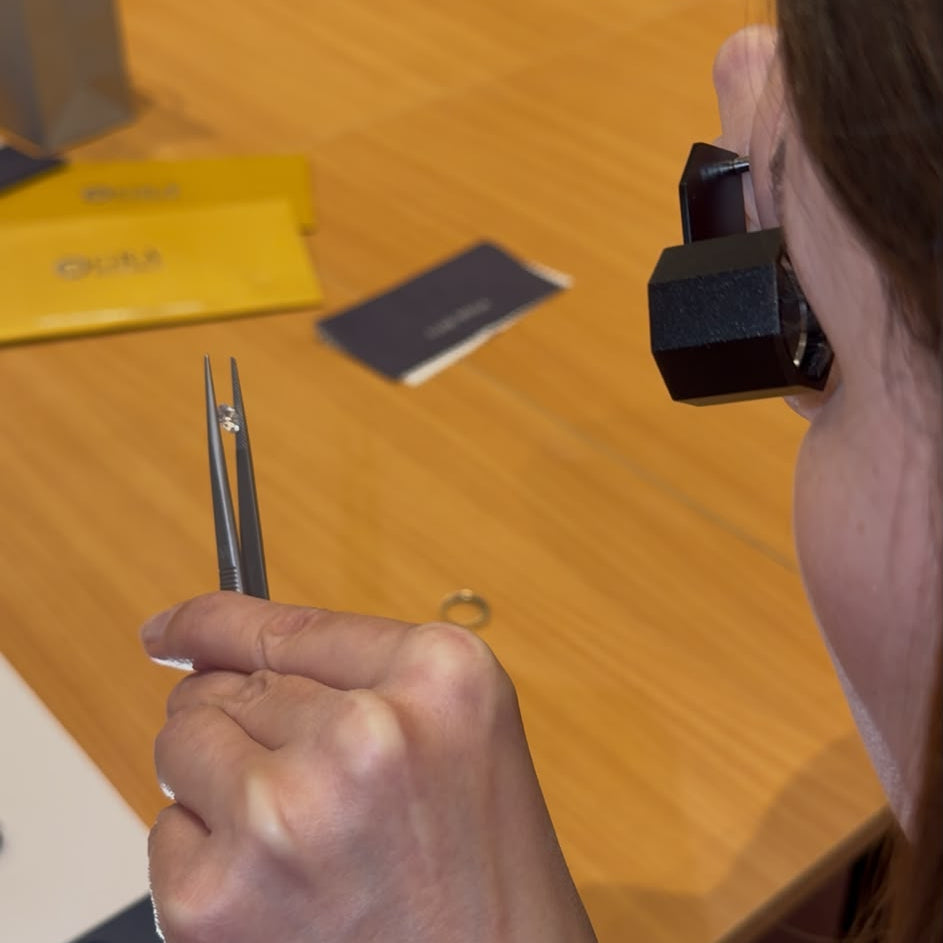 A close-up lifestyle shot of a jeweler using a loupe magnifier and tweezers to examine a ring on a wooden table.
